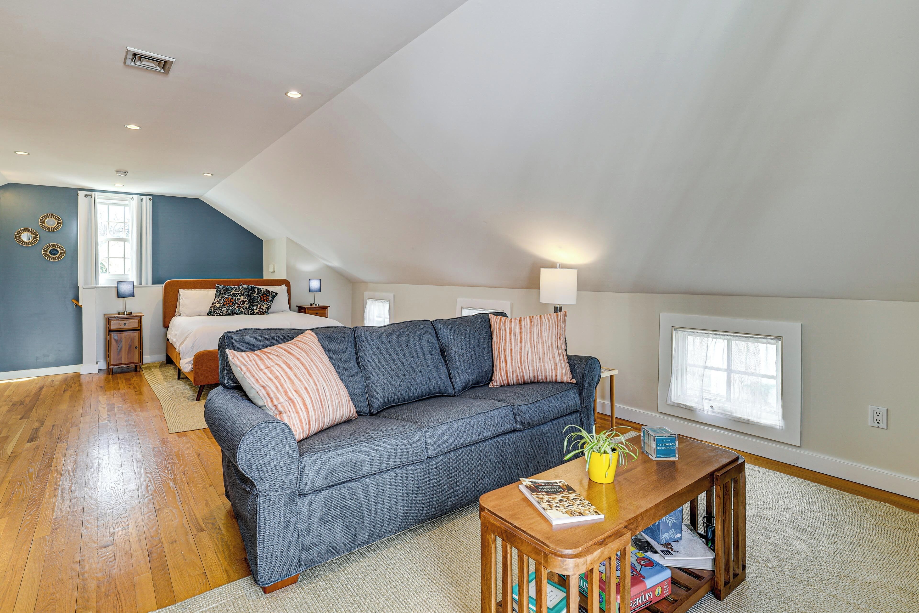 A loft-style bedroom with a bed, a gray sofa, a wooden coffee table, and a window.