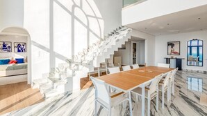 A spacious dining area with a wooden table and white chairs, a white staircase, and a large window providing natural light.