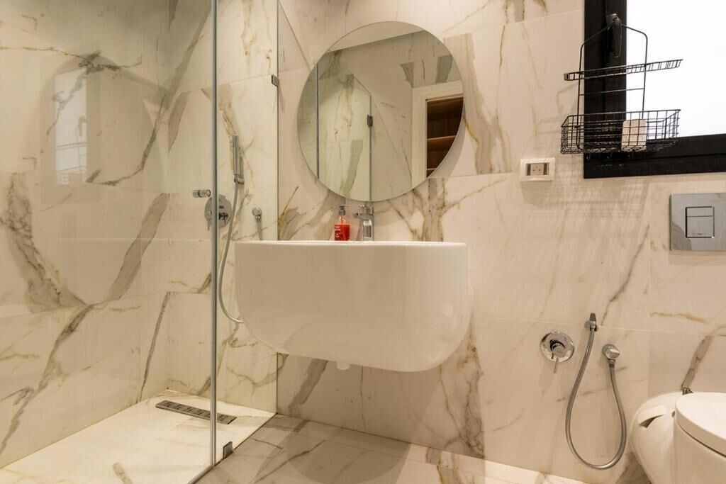 Une salle de bain moderne avec une douche à parois de verre, un mur recouvert de carreaux de marbre, un miroir rond et un lavabo blanc.