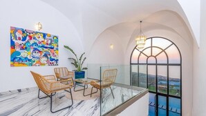 A balcony with a glass railing, a dining area with wicker chairs, a potted plant, and a colorful abstract painting.