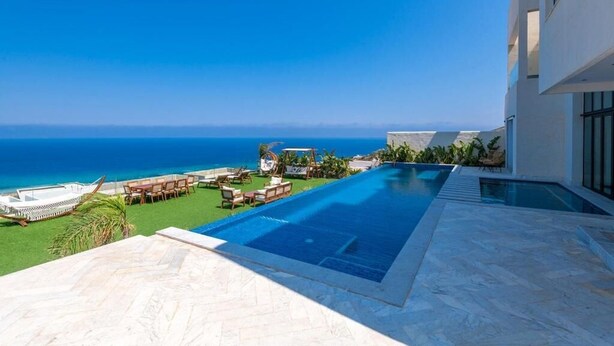 A poolside view with a clear blue sky and ocean, a modern building, and outdoor furniture.
