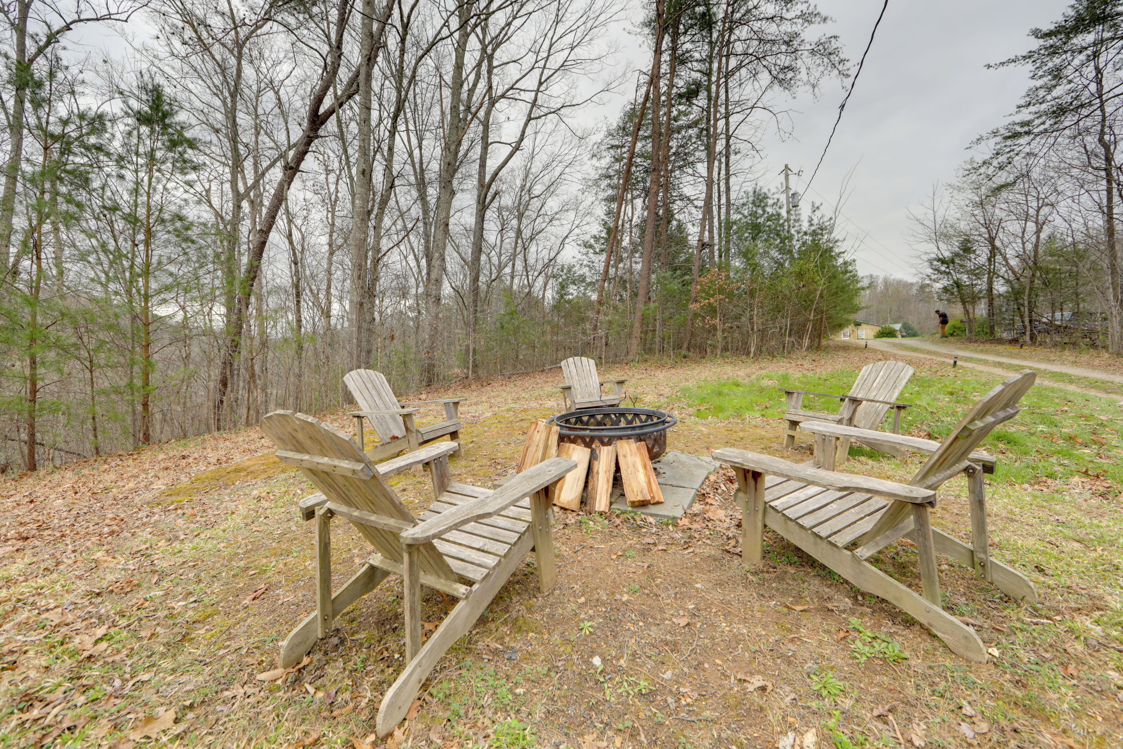 A fire pit with four Adirondack chairs arranged around it in a wooded area.