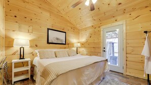 Quarto de cabana de madeira com uma cama, duas luminárias de apoio, um quadro na parede e uma porta para o exterior.