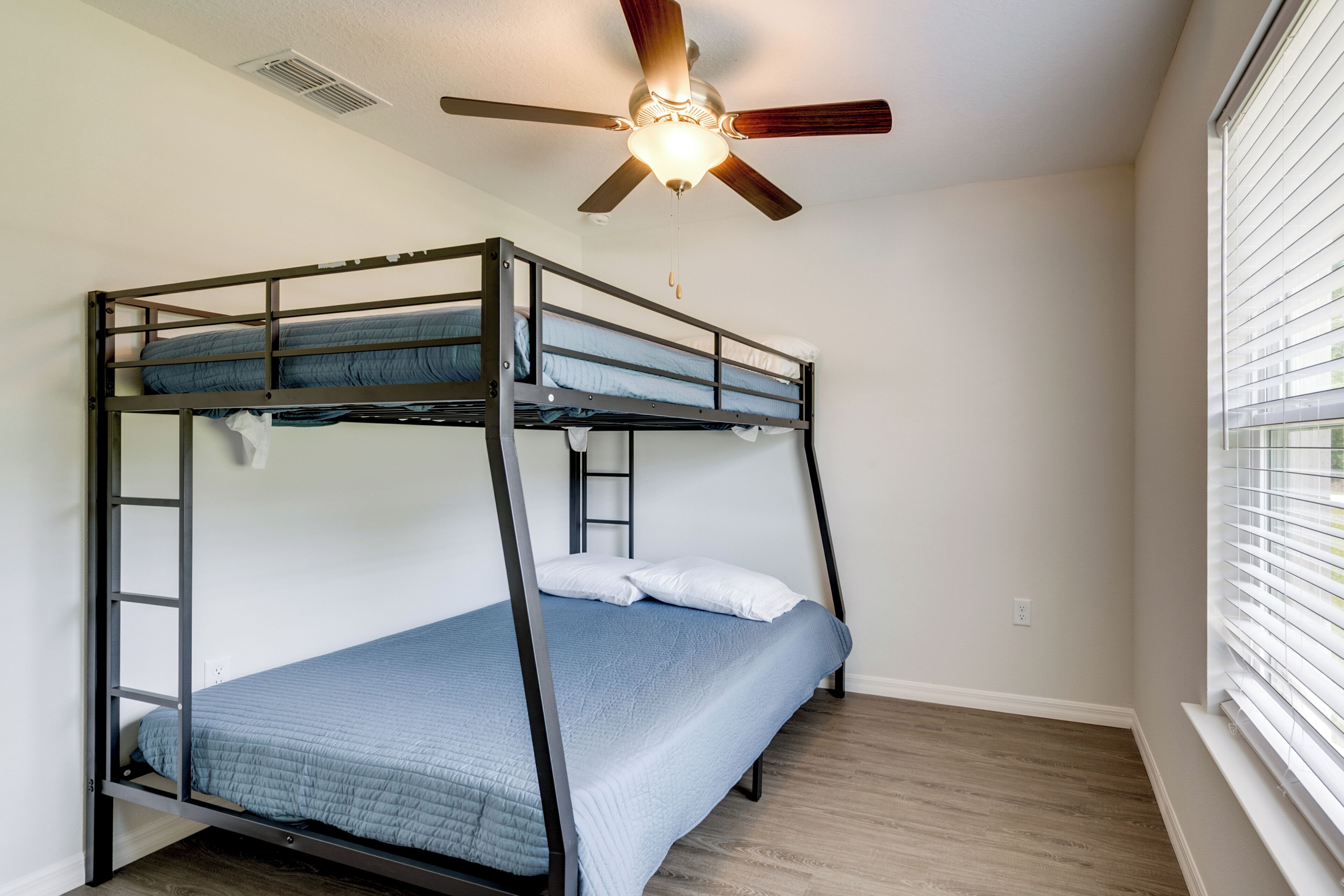A bunk bed with a ceiling fan, wooden flooring, and a window with blinds.