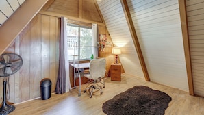Une pièce avec un plafond en pente, un bureau avec une chaise, un ventilateur sur pied, une poubelle et une lampe.