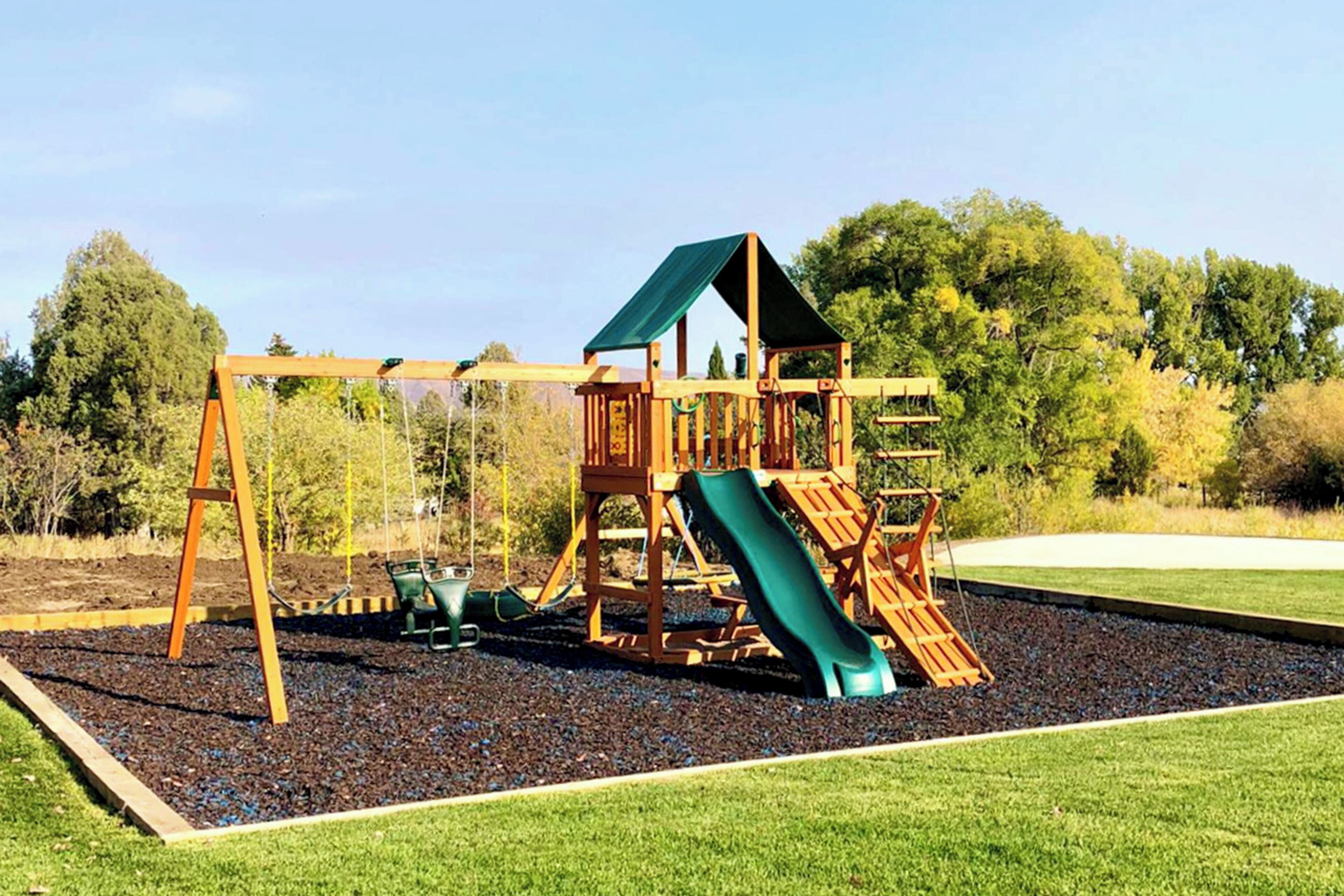 A playground with a wooden slide and swings, surrounded by gravel and grass, with trees in the background.