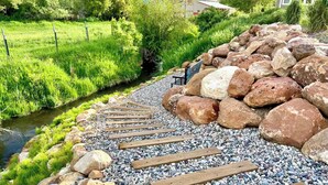 Un sendero de tablones de madera y grandes rocas que conduce a una zona de césped con un pequeño arroyo.
