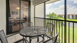 A balcony with a round table and chairs, a sliding glass door leading to a room with a bed and a ceiling fan, and a view of a building and greenery. - Resort-style Fort Myers Gem: Swim, Golf & Cruise! (Fort Myers)
