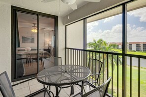 A balcony with a round table and chairs, a sliding glass door leading to a room with a bed and a ceiling fan, and a view of a building and greenery. - Resort-style Fort Myers Gem: Swim, Golf & Cruise! (Fort Myers)