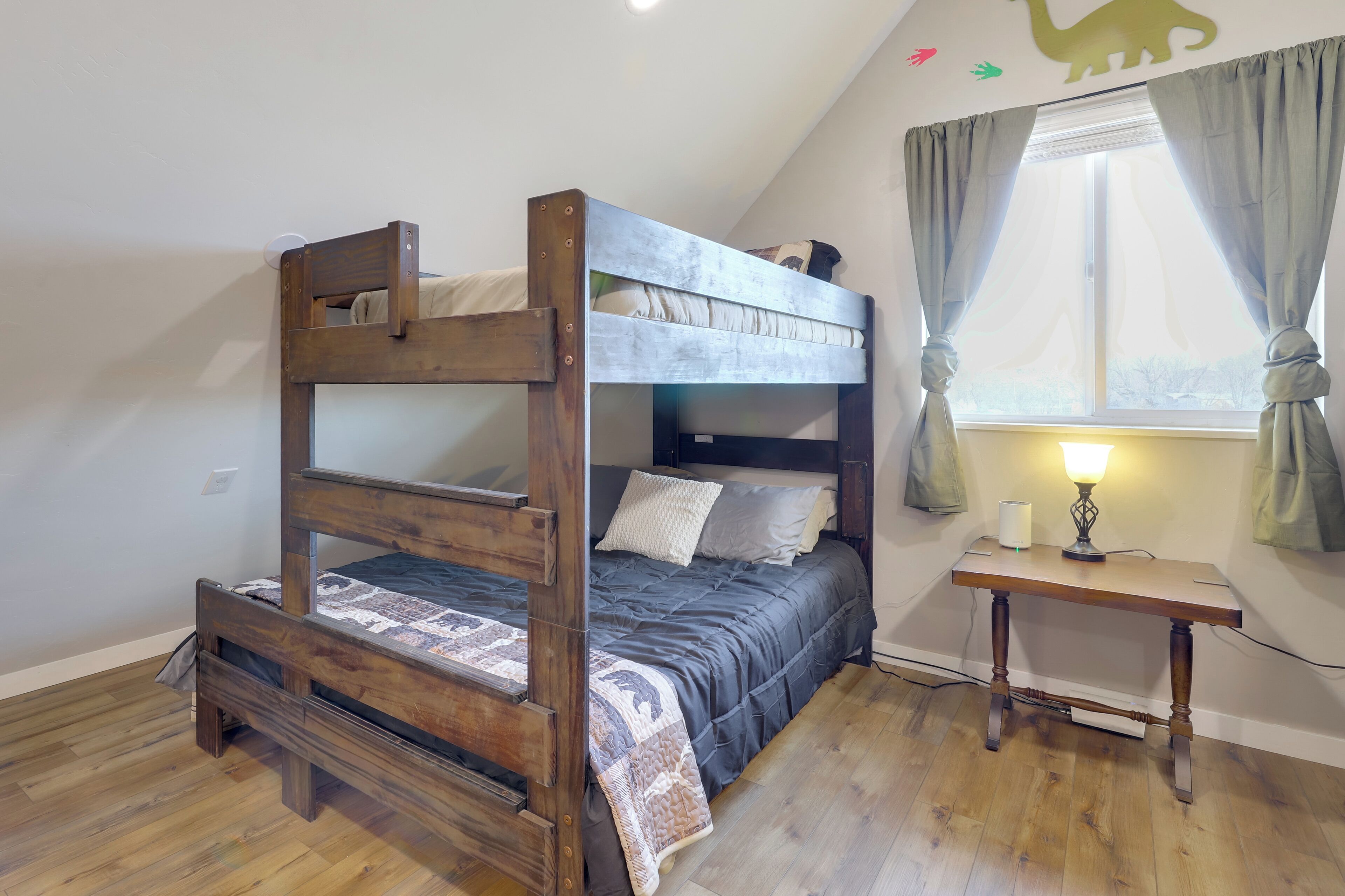 Una cama litera con escritorio y lámpara, suelo de madera y ventana con cortinas.