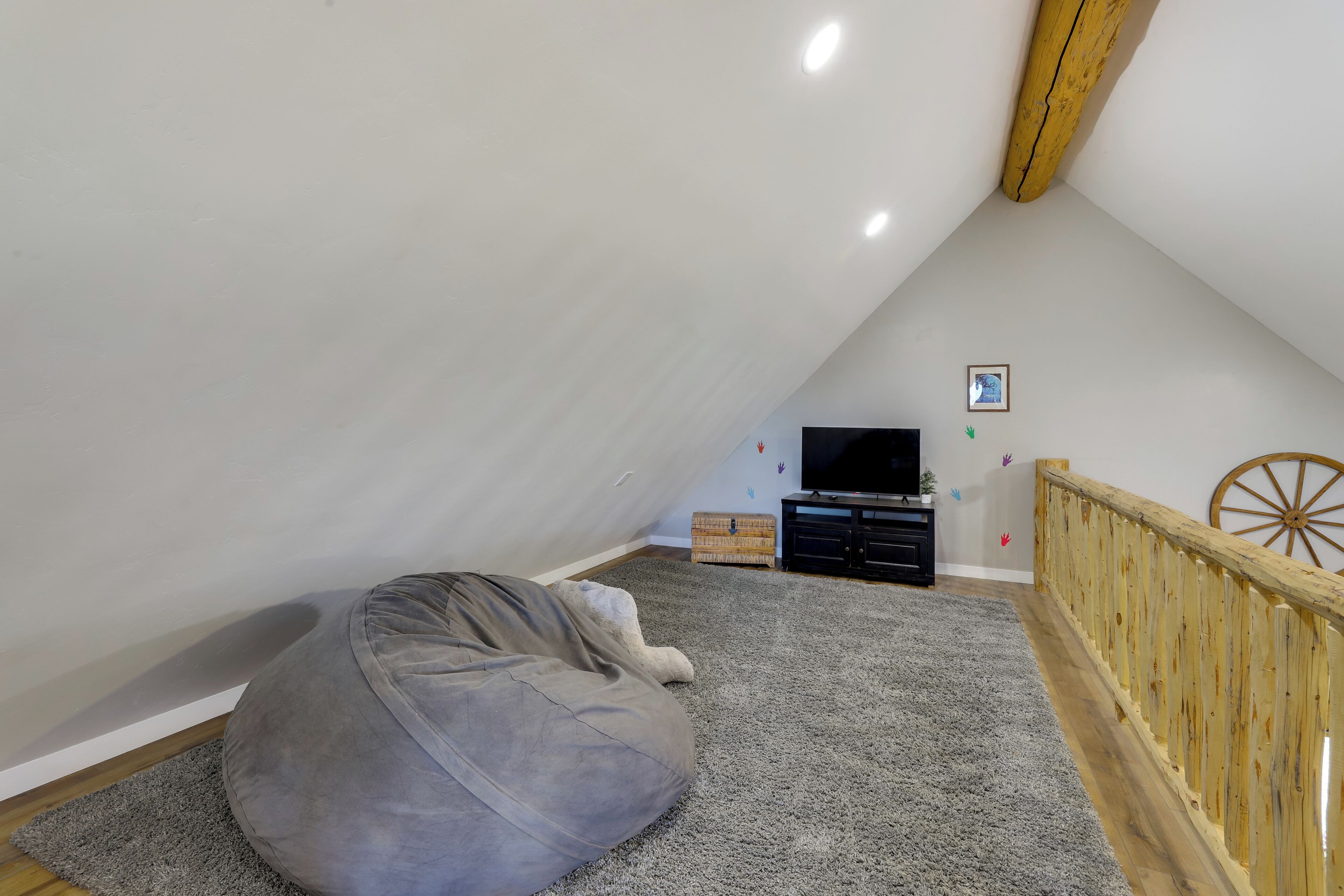 Un dormitorio estilo loft con techo alto, un sillón informal gris, un televisor, una rueda de carro de madera y un cuadro pequeño en la pared.