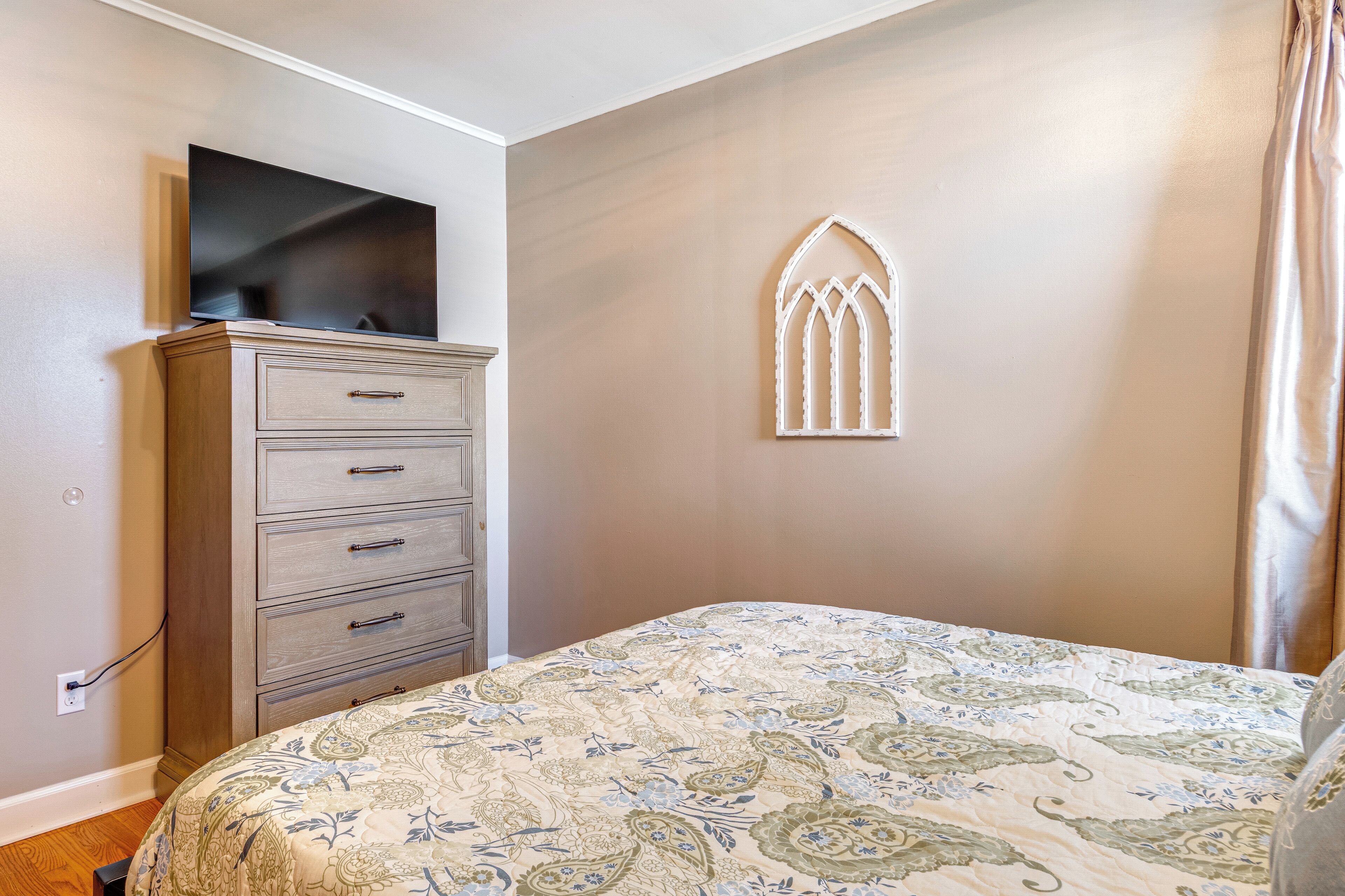 A bedroom with a bed, a dresser, a TV, and a decorative window.