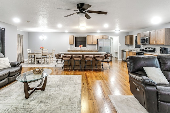 A modern kitchen with a central island, a dining area with a table and chairs, a living room with a sofa and a coffee table, and a ceiling fan.