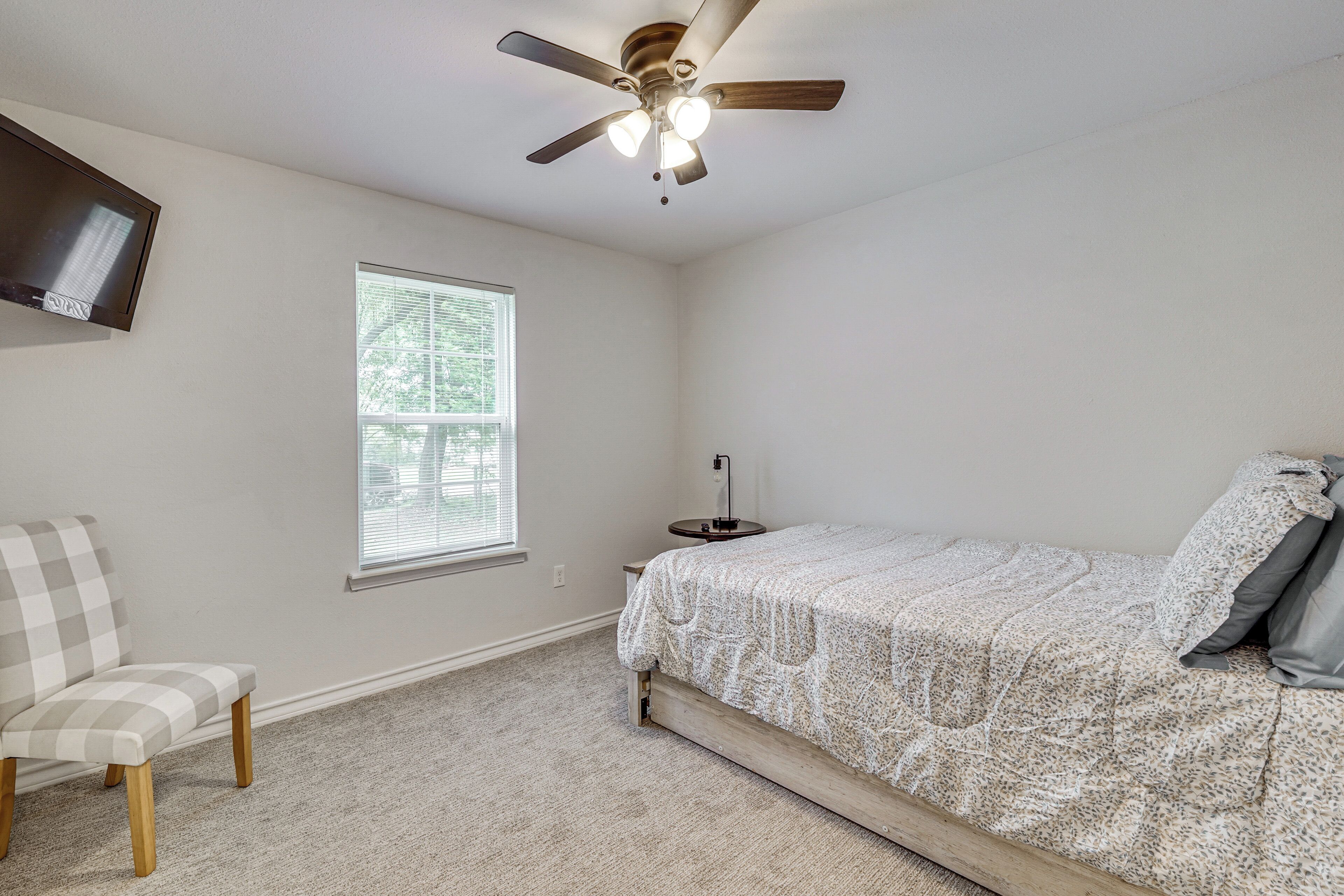 A bedroom with a bed, a chair, a ceiling fan, a TV, and a window.