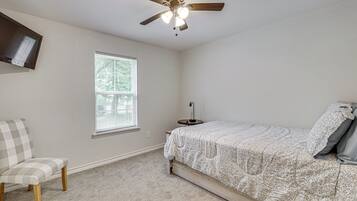 A bedroom with a bed, a chair, a ceiling fan, a TV, and a window.