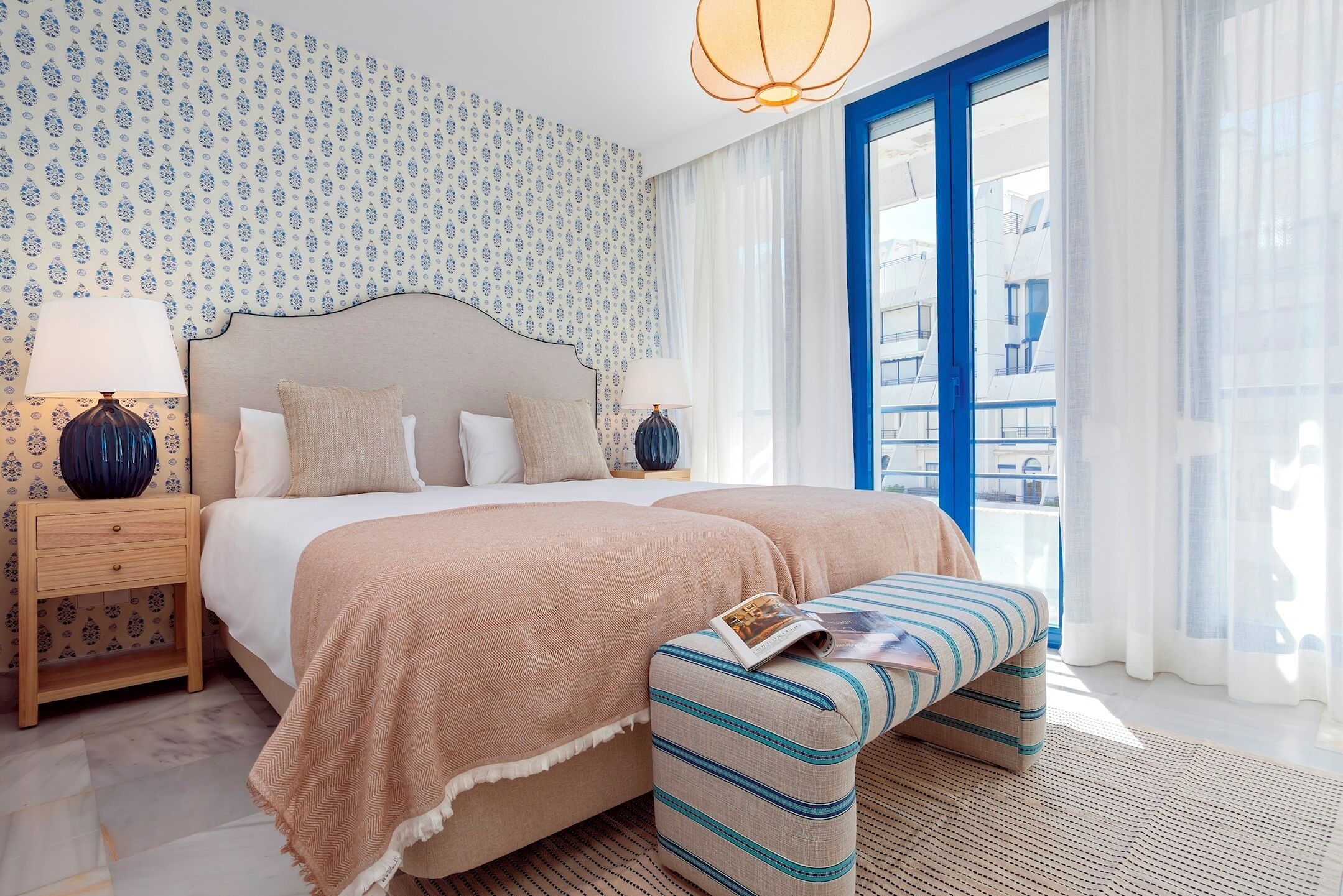 Quarto de hotel com cama, mesas de cabeceira, um banquinho, janela com cortinas e papel de parede estampado.