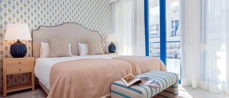 Quarto de hotel com cama, mesas de cabeceira, um banquinho, janela com cortinas e papel de parede estampado.