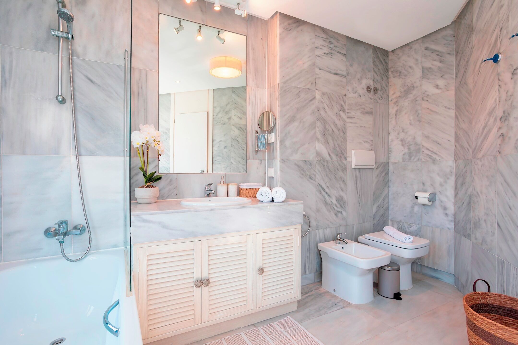 Een moderne badkamer met een marmeren wand, een grote spiegel, een ligbad, een wastafelmeubel en twee toiletten.