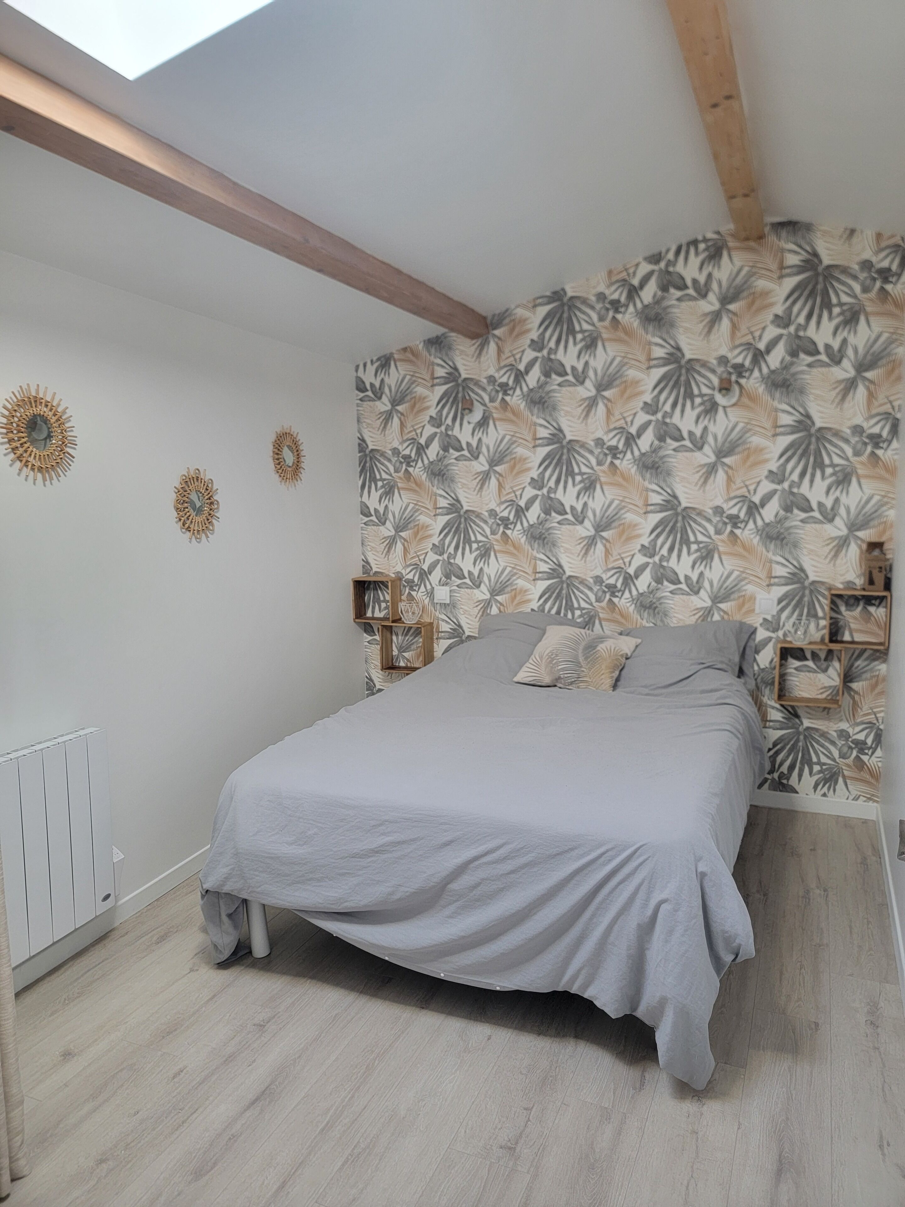 Un dormitorio con techo inclinado, una cama con cubrecama gris, una pared con estampado de hojas tropicales y una viga de madera.