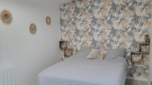 Een slaapkamer met schuine plafond, een bed met grijs beddegoed, een muur met een tropisch bladpatroon, en een houten balk.