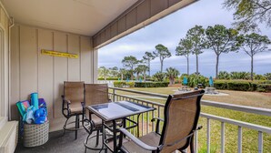 A covered outdoor seating area with wicker chairs and a table, overlooking a landscaped garden with trees and a view of the ocean. - Sunset Rentals | Ocean Dunes 121 (Hilton Head Island)