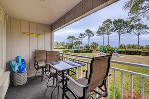 A covered outdoor seating area with wicker chairs and a table, overlooking a landscaped garden with trees and a view of the ocean. - Sunset Rentals | Ocean Dunes 121 (Hilton Head Island)