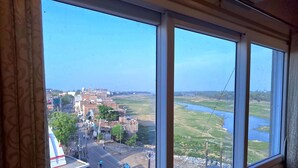 Une vue à travers une fenêtre ornée d’un rideau à motifs, un ciel dégagé et une rivière avec un pont visible au loin.