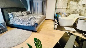 Un dormitorio moderno con una cama grande, una zona de estar separada, un baño con paredes de vidrio y una bañera, y un tocador con espejo.