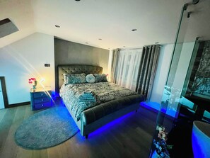 Un dormitorio moderno con cabecera capitonada, cama con ropa de cama estampada, mesita de noche con lámpara y separador de vidrio.