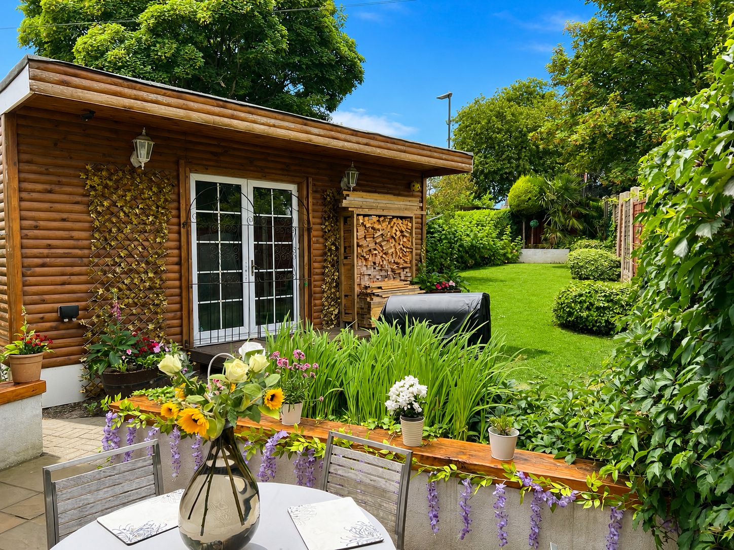 A wooden cabin with a large window, a barbecue grill, and a garden with various plants and flowers.