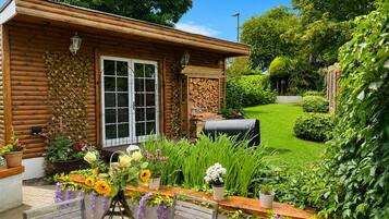 A wooden cabin with a large window, a barbecue grill, and a garden with various plants and flowers.