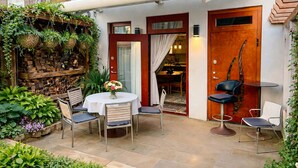 Un patio con una mesa redonda y sillas, una puerta de madera y un estante en la pared con plantas.