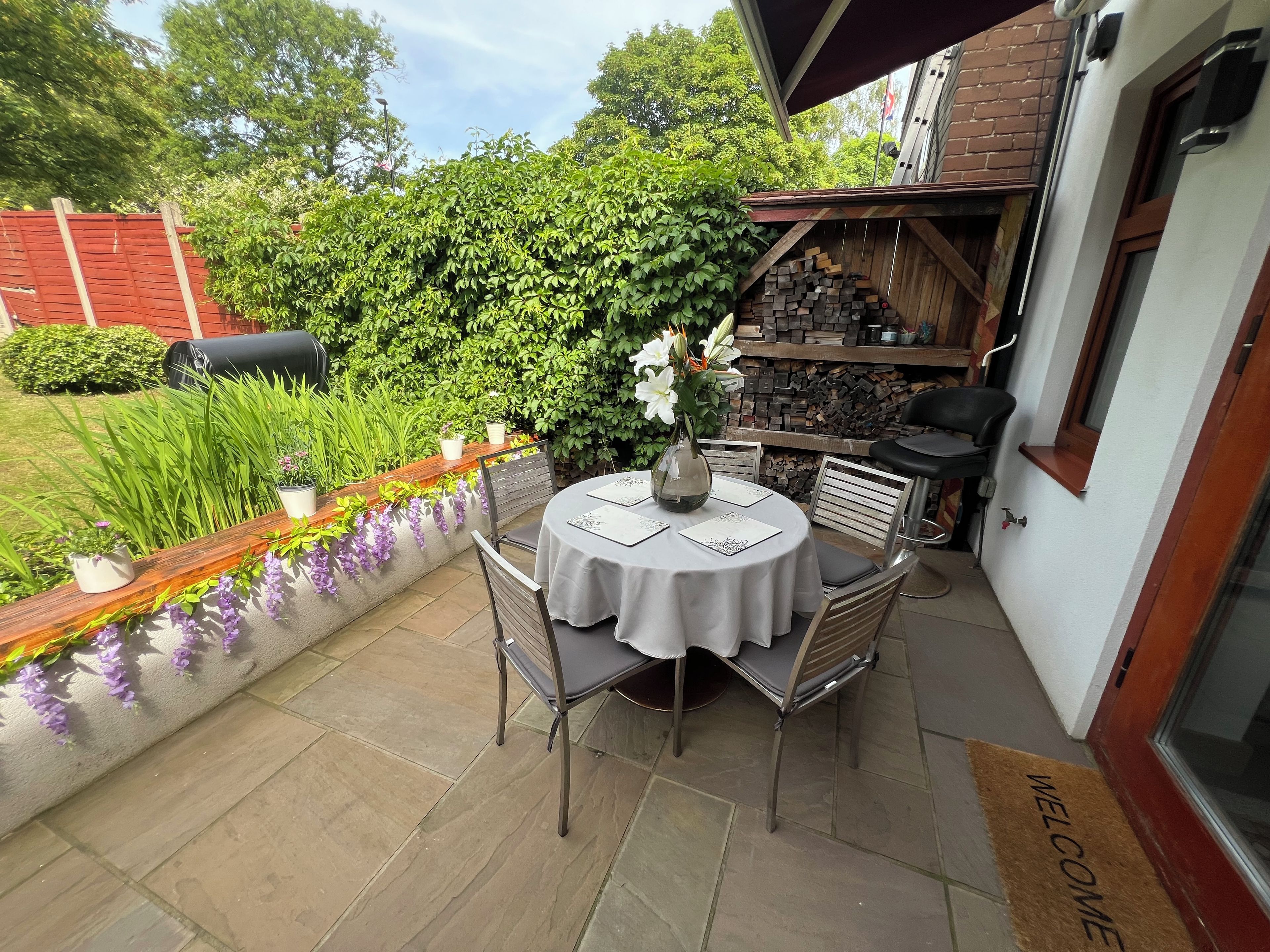A patio with a round table, chairs, and a vase with flowers. There is a brick wall and a wooden fence in the background.