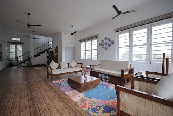 A spacious living area with a wooden floor, a sofa, a coffee table, and a rattan chair. There are ceiling fans, a staircase, and large windows.