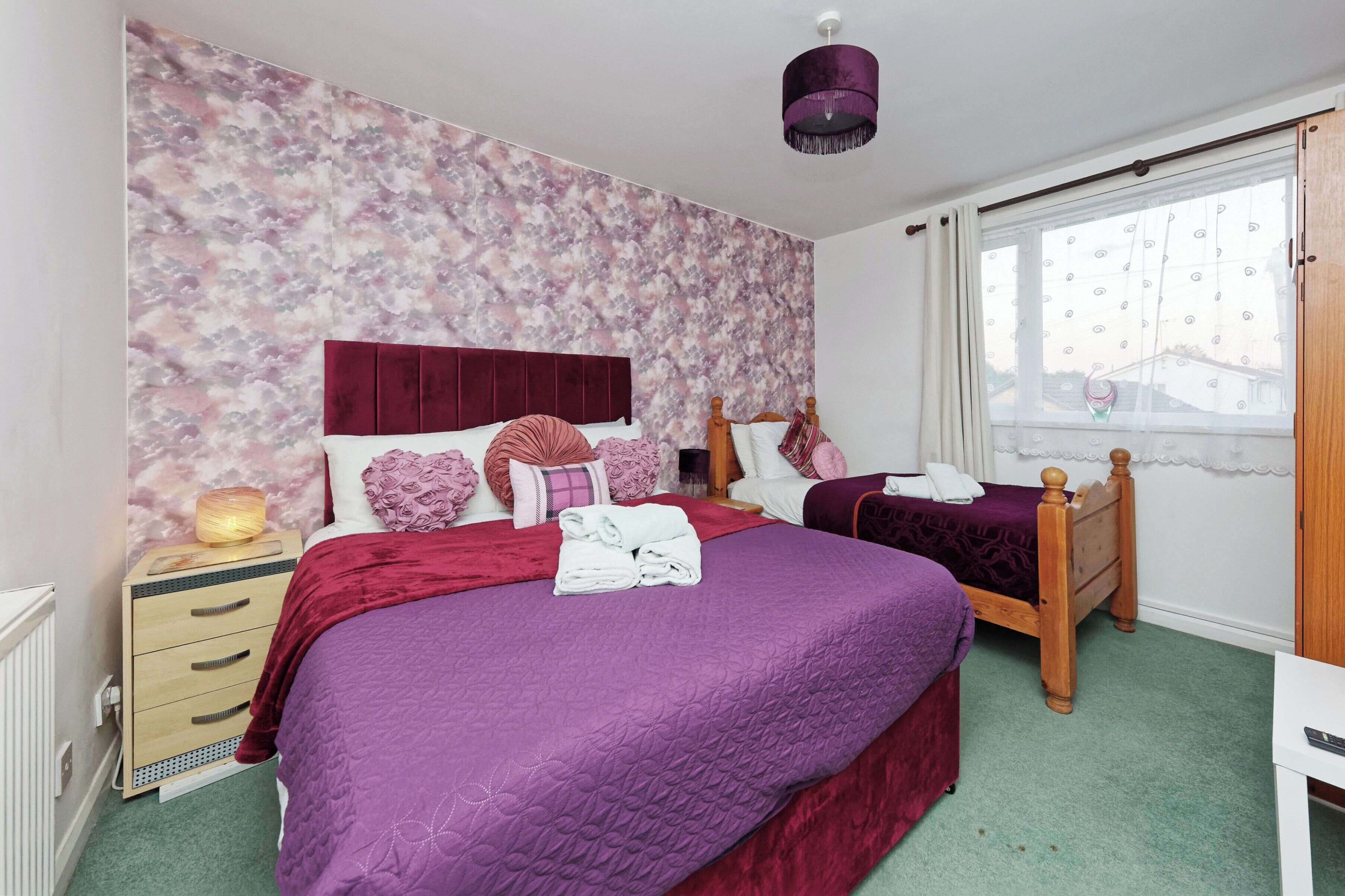 Une chambre avec deux lits, une table de chevet, une lampe, une fenêtre avec des rideaux et un luminaire au plafond.