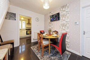 Un coin repas avec une table ronde, des chaises, une horloge, un téléviseur et, au fond, une cuisine.