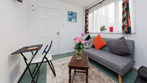 Ein Wohnzimmer mit einer Couch, einem kleinen Tisch, einem Sessel, einem Fernseher, einer Tür, einem Fenster mit Vorhängen und einer Pflanze.