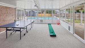 Ein überdachter Innenbereich mit einem Tischtennisplatte, einer Shuffleboard-Bahn und Blick auf einen Garten mit Pool.