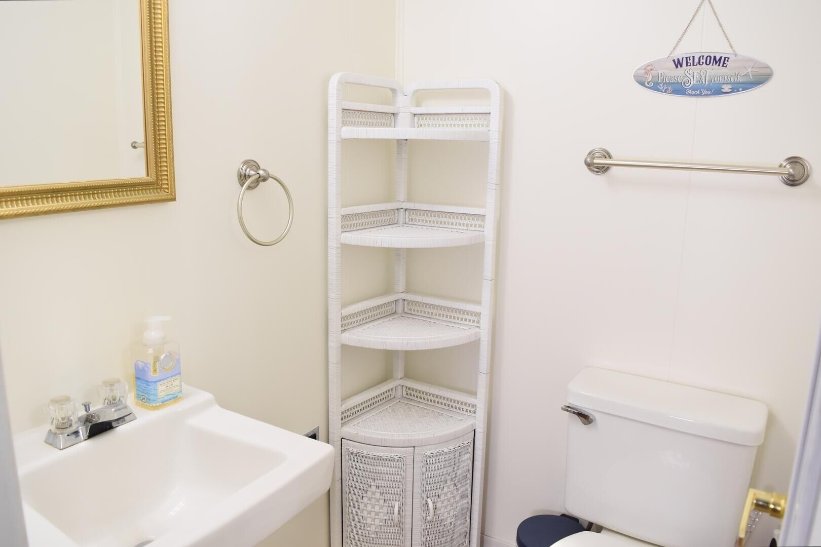 A bathroom with a white sink, a toilet, a wicker shelf unit, and a welcome sign.