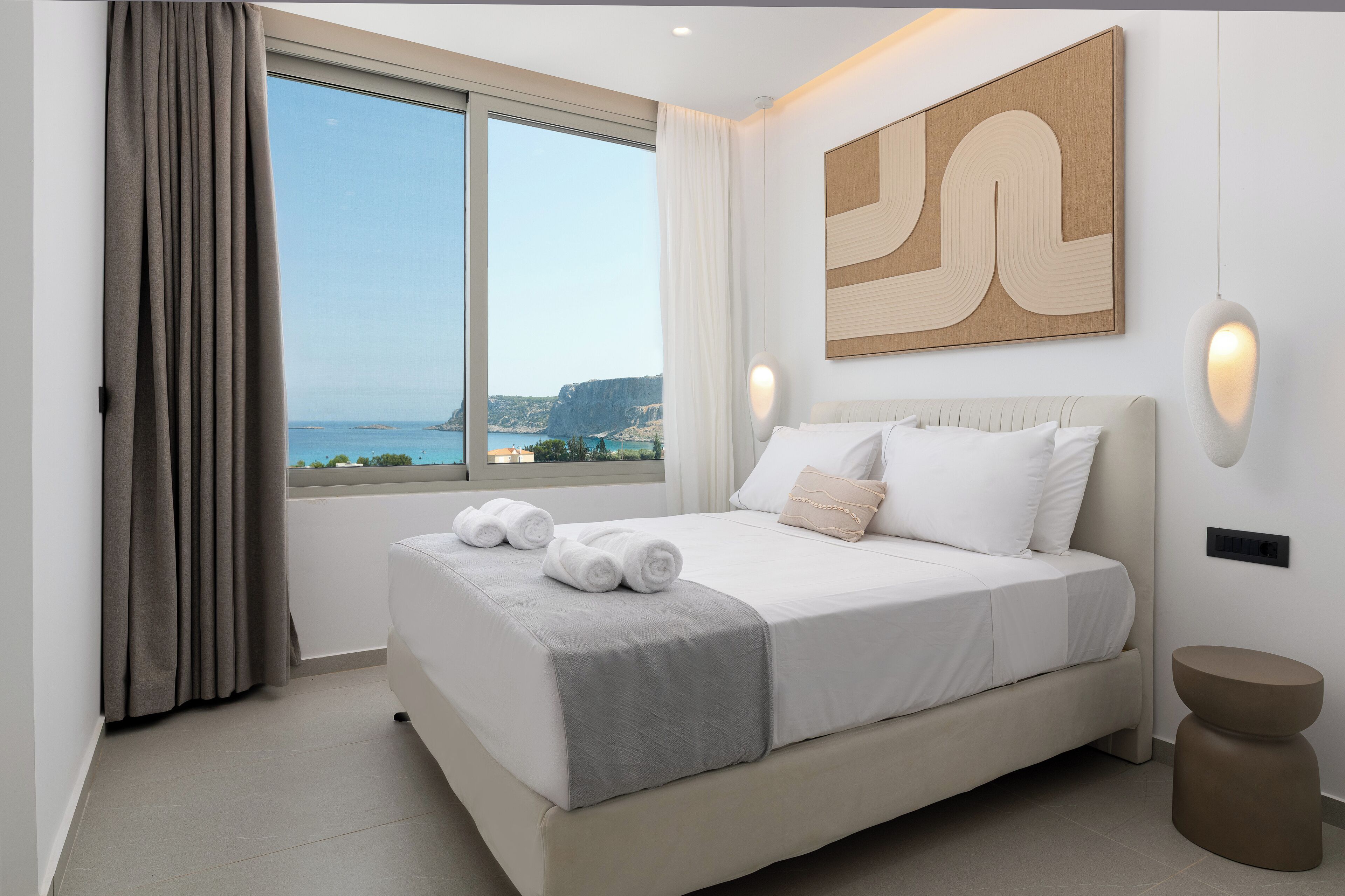 Un dormitorio con un ventanal grande, una cama con ropa de cama blanca y vista al mar, además de un cuadro abstracto moderno en la pared.