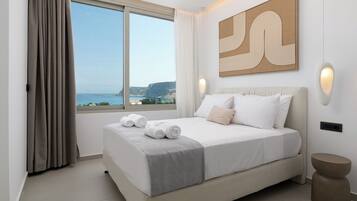 Un dormitorio con un ventanal grande, una cama con ropa de cama blanca y vista al mar, además de un cuadro abstracto moderno en la pared.