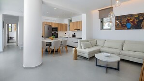 Ein modernes Wohnzimmer mit einer Couch, einem Esstisch und Stühlen. Im Hintergrund befindet sich eine Küche mit Holzschränken und einem Kühlschrank.