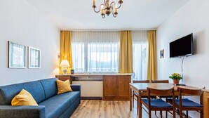 A modern living room with a sofa, a dining table with chairs, a TV mounted on the wall, and a kitchen area with a window. - Grimmingblick Apartment with Pool and Spacious Terrace (Sonnenalm)