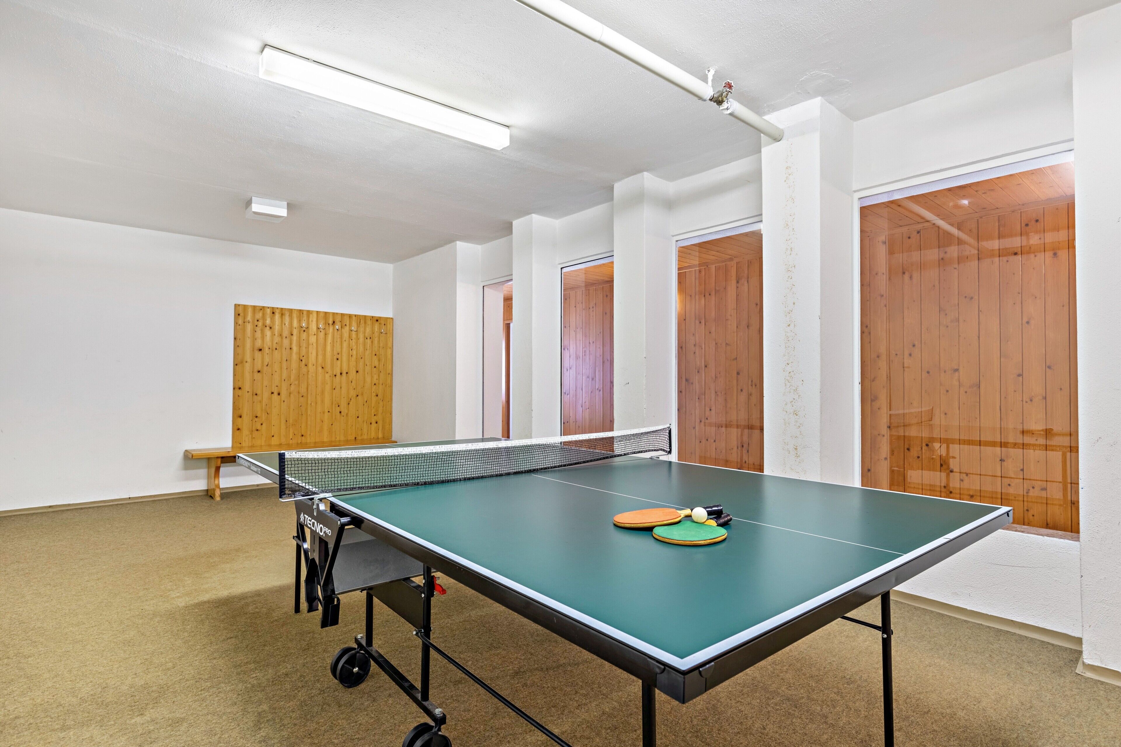 Una mesa de ping pong en una habitación con paredes blancas, paneles de madera y un banco.