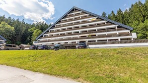 A multi-story hotel with balconies, a parking area, and a grassy hill in the foreground. - Grimmingblick Apartment with Pool and Spacious Terrace (Sonnenalm)