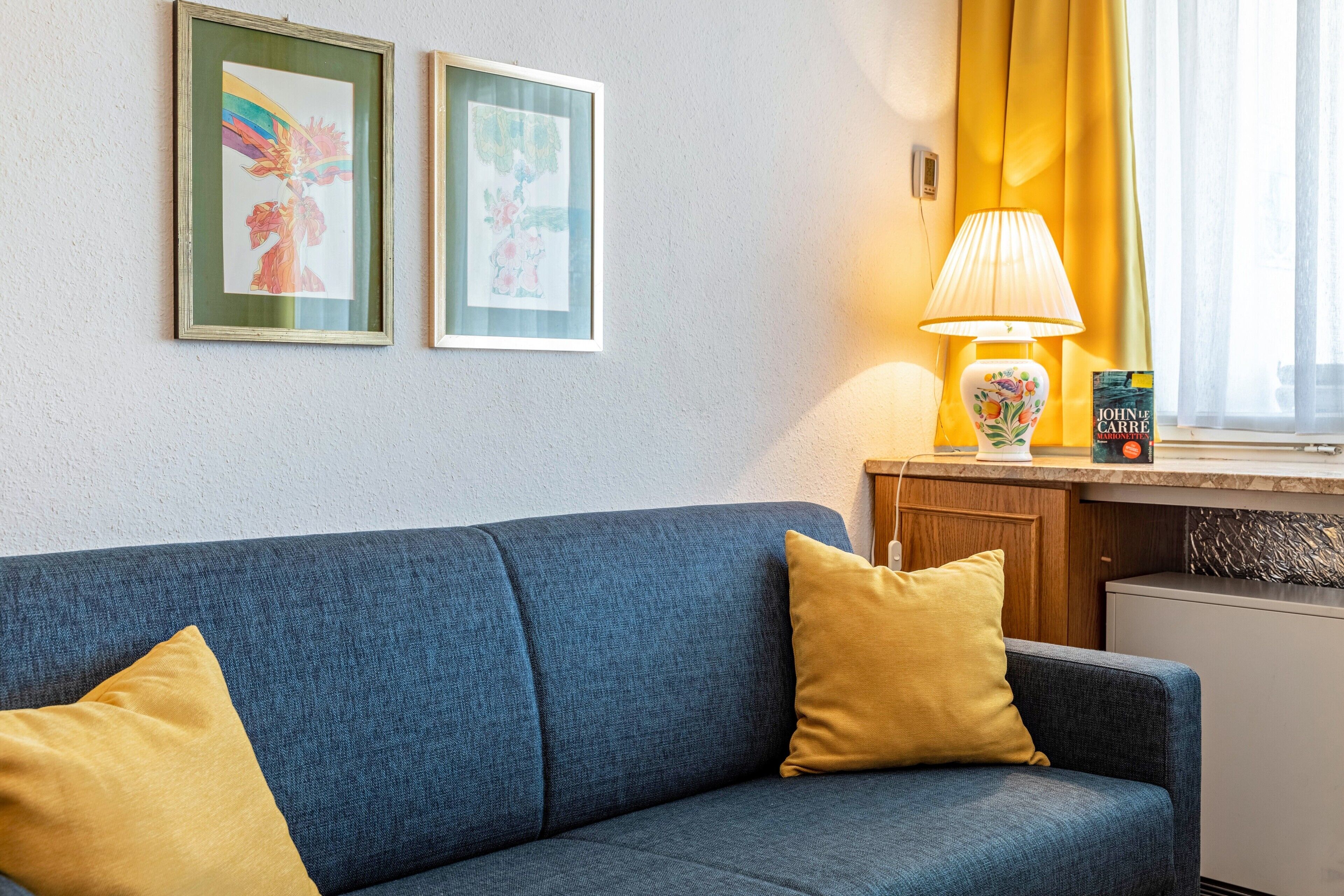 A room with a blue sofa, two yellow pillows, a lamp, a small table with a book, and two framed pictures on the wall.