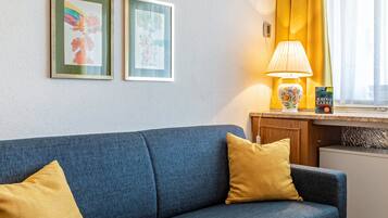 A room with a blue sofa, two yellow pillows, a lamp, a small table with a book, and two framed pictures on the wall.