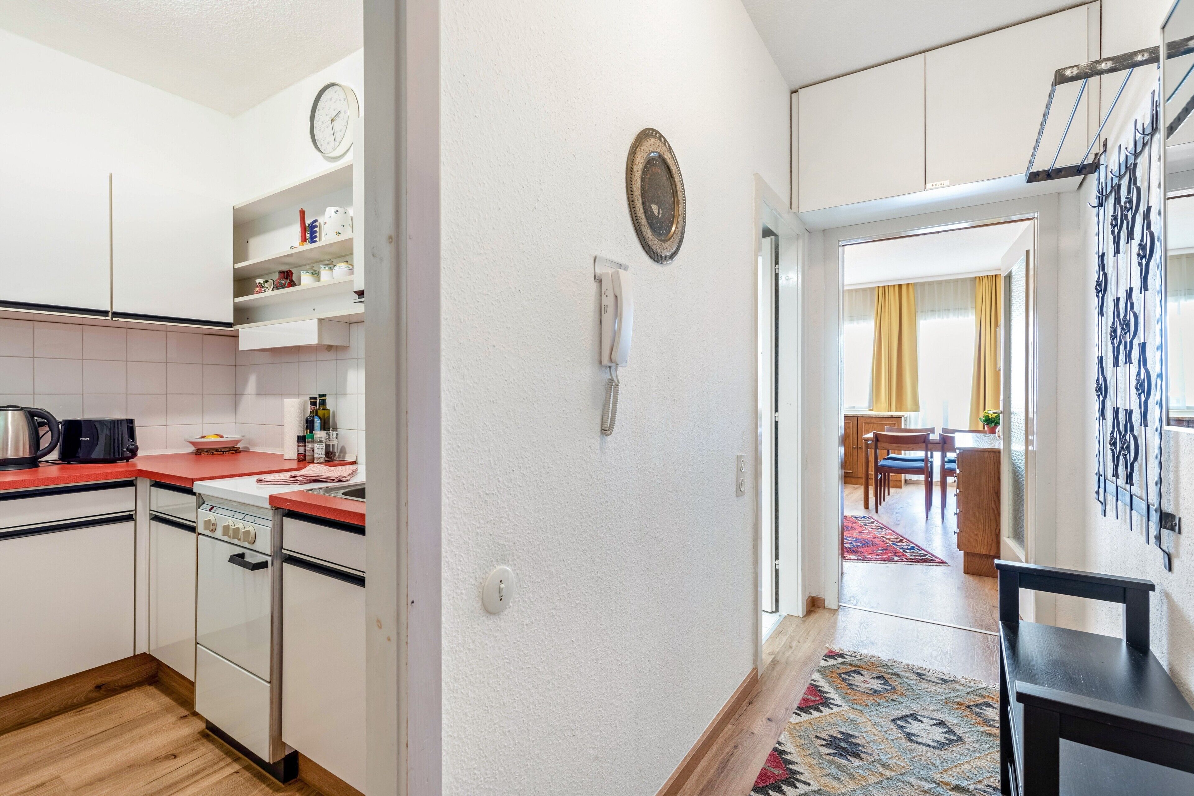 Une cuisine avec des meubles blancs, un plan de travail rouge, une horloge, un téléphone et un tapis à motifs qui mène à un coin repas.