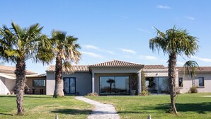 Een huis met een houten steiger die uitkomt op een waterpartij, omgeven door palmbomen en een helderblauwe lucht.