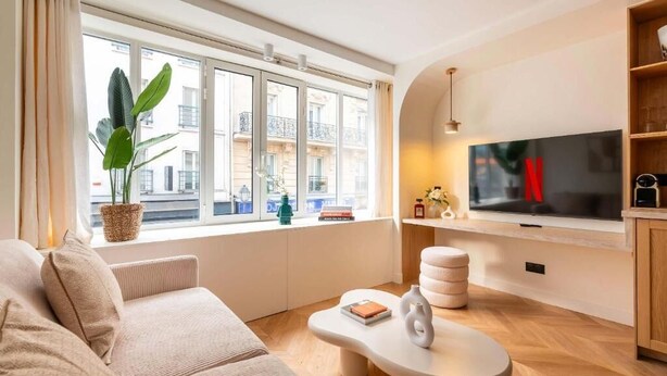 Ein modernes Wohnzimmer mit einer Couch, einem Couchtisch, einem Fernseher und einem großen Fenster mit Blick auf Gebäude.